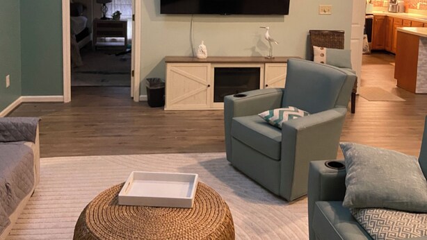 A living room with a flat-screen TV, a ceiling fan, a sofa, a coffee table, and a kitchen in the background. - Quiet Calabash neighborhood convenient to everything (Calabash)