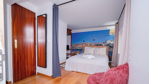 A bedroom with a cityscape view, a wooden door, a bed with white bedding, and a pink chair. - 1 Bedroom. 1 Washroom. 1 Living Room. 1 Double bed, 1 Sofa bed Apartment (Lisbon)