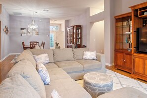 Ein Wohnzimmer mit Ecksofa, Hocker, Holzschrank und einer Deckenleuchte.