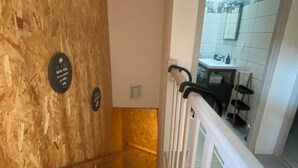 Un escalier avec des murs en bois et une rampe blanche, menant à une salle de bain dont un mur est recouvert de carreaux et qui est équipée d’un lavabo.
