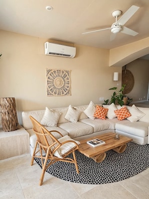 A living room with a white sofa, a wooden coffee table, a wicker chair, a black and white patterned rug, a ceiling fan, and a wall-mounted air conditioner. - Oceanview Suite Perfect Hideaway for Two (La Majahua)