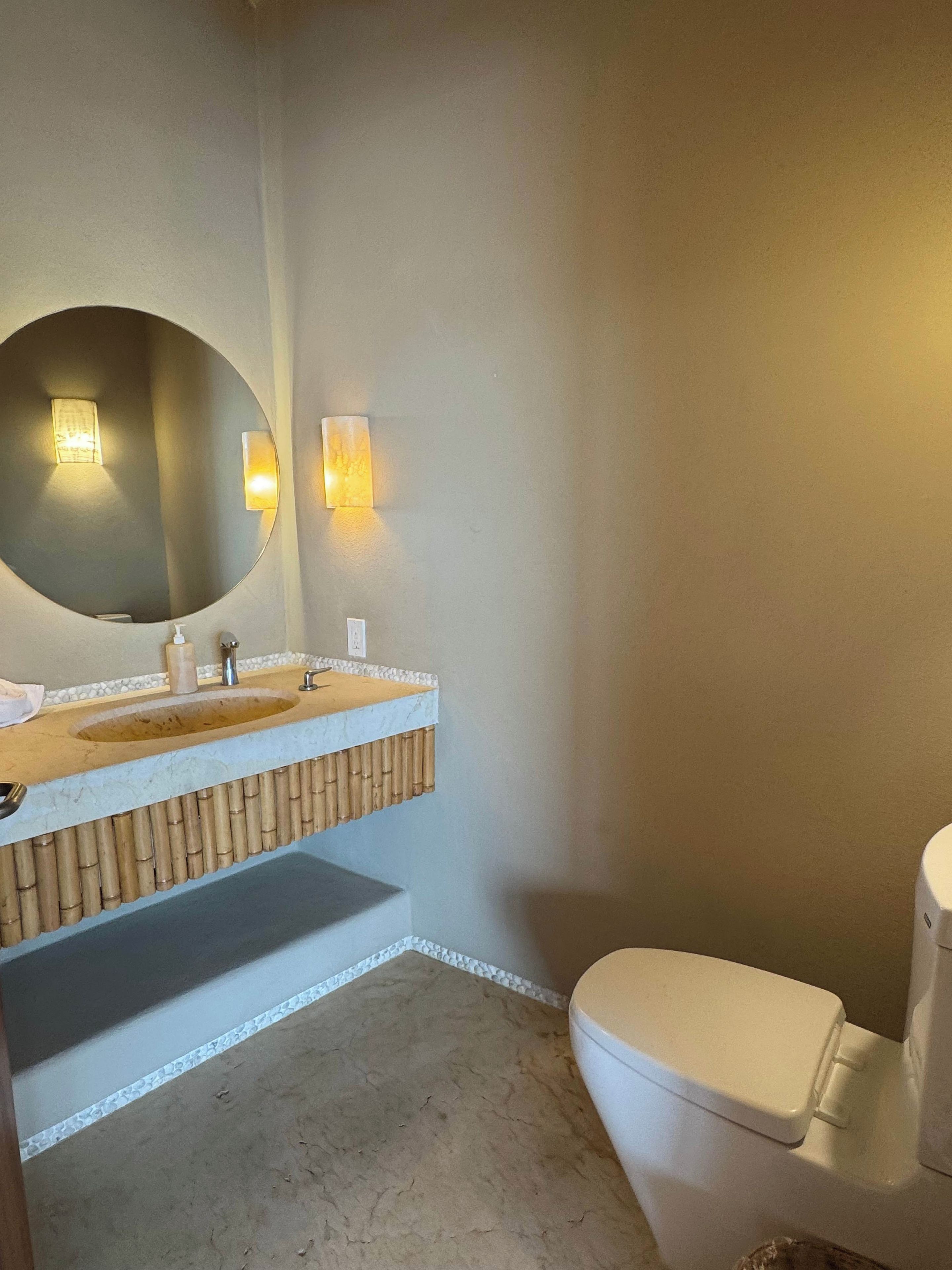 A bathroom with a round mirror, a sink with a marble countertop, two wall-mounted lights, and a toilet.