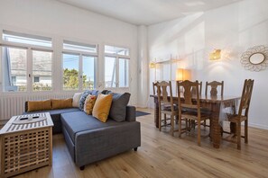 Una sala de estar con un sofá gris, una mesa de comedor de madera y sillas. Hay ventanas grandes con marcos blancos, un radiador y luces fijadas a la pared.