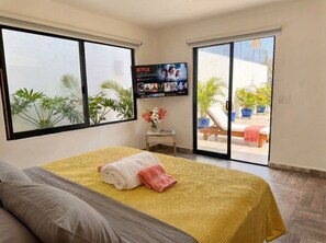 Ein Hotelzimmer mit einem Bett, einem an der Wand montierten Fernseher, einem kleinen Tisch mit einer Vase und einer Glastür, die zu einem Außenbereich mit Blumentöpfen führt.