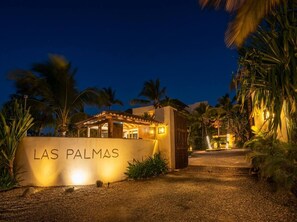 Un bâtiment sur lequel on peut lire l’inscription « LAS PALMAS », illuminé la nuit, entouré de palmiers et bordé d’un chemin de gravier.