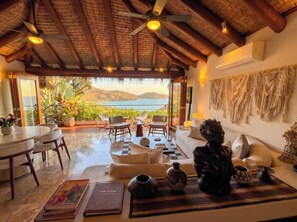 A room with a view of the sea, a ceiling fan, a sofa, a table with books, and decorative items.
