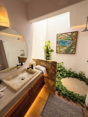 A modern bathroom with a large mirror, two sinks, a shower, a stone accent wall, a potted plant, and a painting.