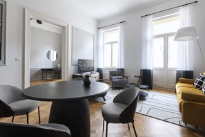 Un salon moderne avec une table ronde, des fauteuils gris, un canapé jaune et une grande fenêtre avec des rideaux.