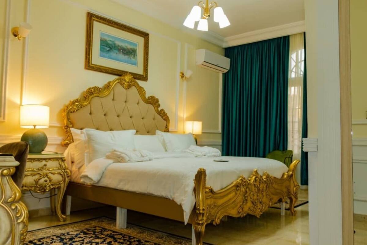 A hotel room with a large bed, ornate headboard, two bedside lamps, a framed picture on the wall, and a window with curtains.