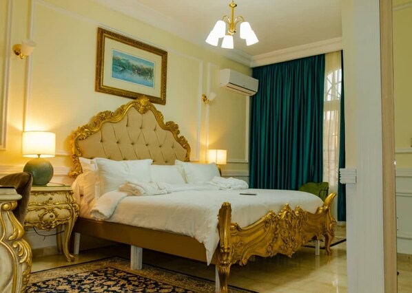 A hotel room with a large bed, ornate headboard, two bedside lamps, a framed picture on the wall, and a window with curtains. - Hôtel Chateau Blanc  (Yamoussoukro)