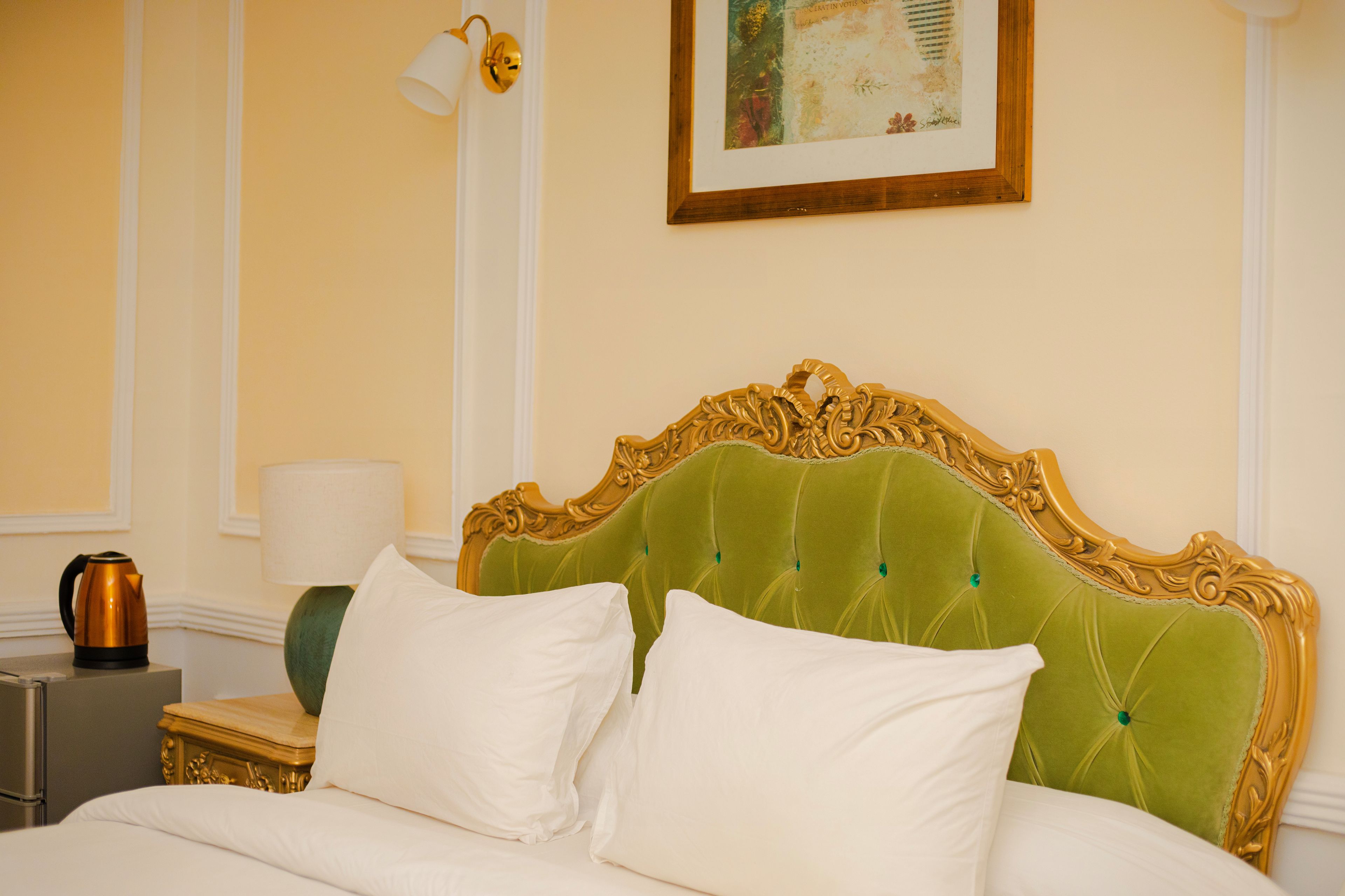 Une chambre d’hôtel avec une tête de lit recouverte d’un tissu vert, du linge de lit blanc, une table de chevet avec une lampe et une œuvre d’art encadrée accrochée au mur.