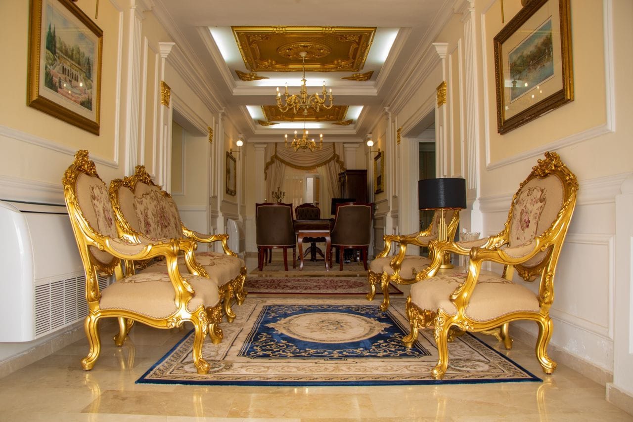 Um amplo salão com cadeiras douradas ricamente decoradas, um candelabro e obras de arte emolduradas nas paredes.