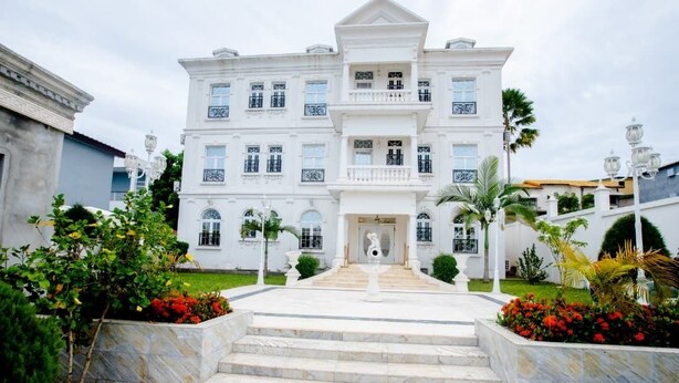 A large, multi-story building with a grand entrance, steps leading up to it, and a well-maintained garden with various plants and flowers. - Hôtel Chateau Blanc  (Yamoussoukro)