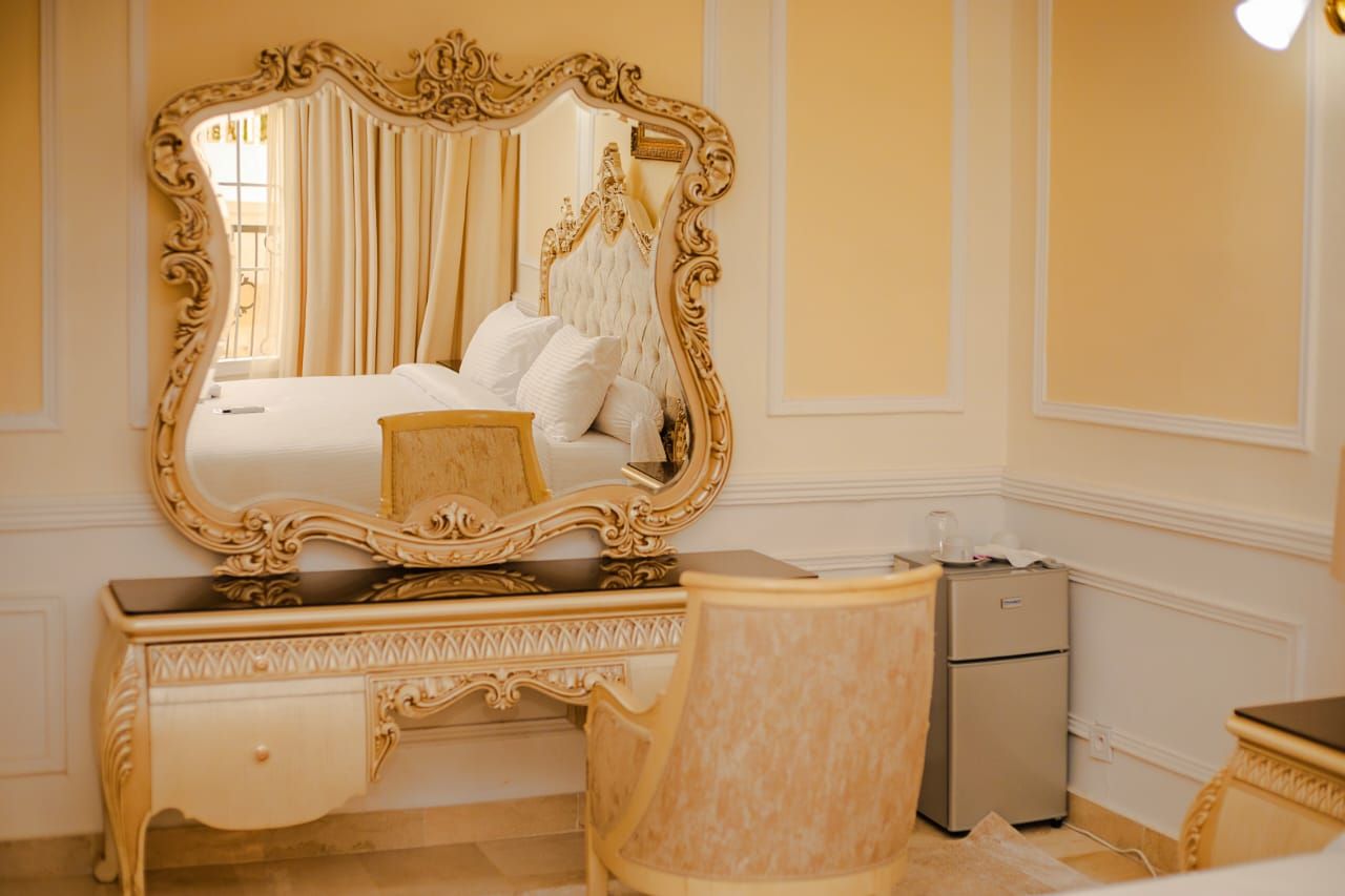 A bedroom with a large ornate mirror, a bed with white linens, a vanity with a mirror, and a small refrigerator.