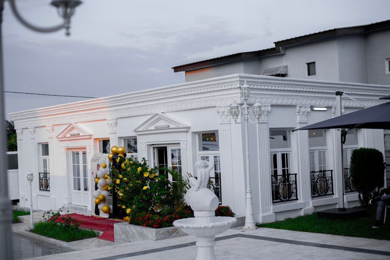 Um edifício branco com um tapete vermelho que leva à entrada, uma fonte e um jardim bem cuidado.
