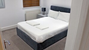 A neatly made bed with white linens and two pillows, a bedside lamp, a radiator, and a window with blinds. - Comfortable, modern 2 bed apartment in the heart of the Old Town (Hemel Hempstead)