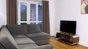 A living room with a grey sectional sofa, a flat-screen TV on a wooden stand, and a window with curtains. - Comfortable, modern 2 bed apartment in the heart of the Old Town (Hemel Hempstead)