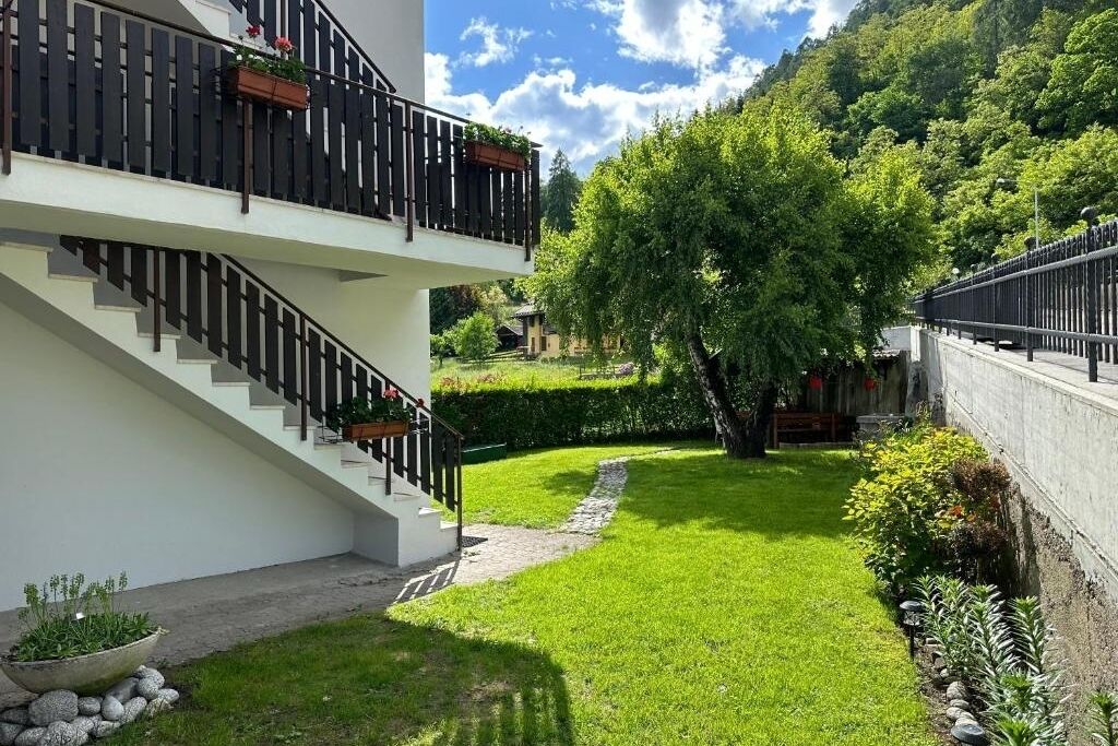Un bâtiment moderne à deux étages, doté d’une façade blanche, d’un escalier avec des rampes noires, d’un jardin bien entretenu avec un sentier et d’un grand arbre.