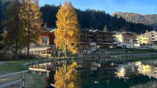 Hotel aan een meer, met een spiegelbeeld in het water, omgeven door bomen en bergen.