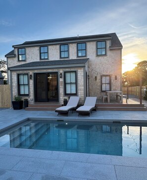 A two-story house with a swimming pool, two lounge chairs, and a wooden deck.