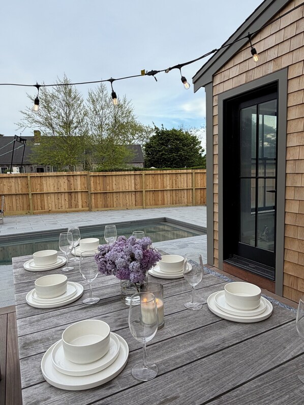 A wooden deck with a dining table set for six, surrounded by a wooden fence, and a building with large windows in the background.