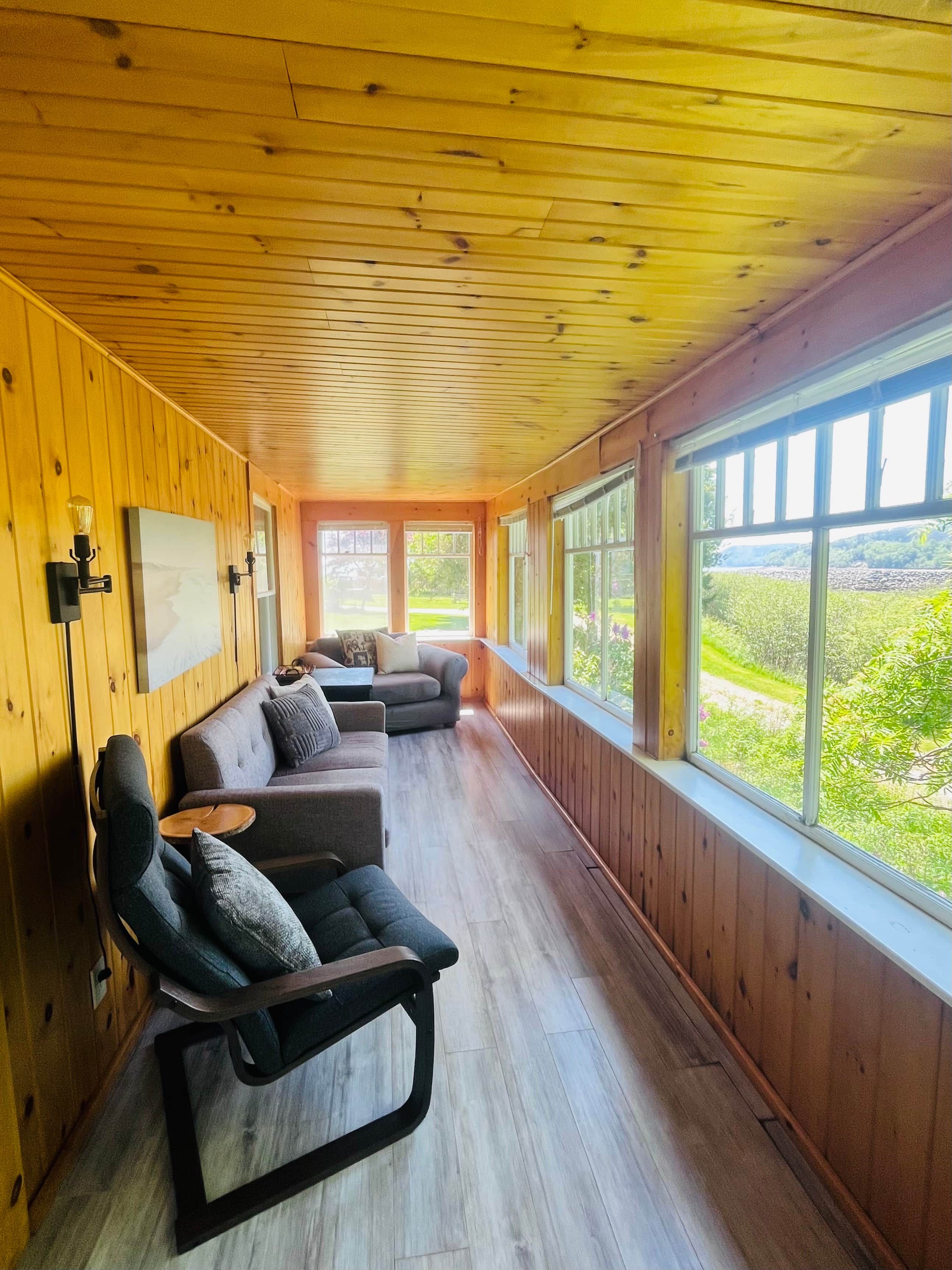 Une pièce aux murs et au plafond en bois, avec une grande fenêtre et un canapé agrémenté de coussins.