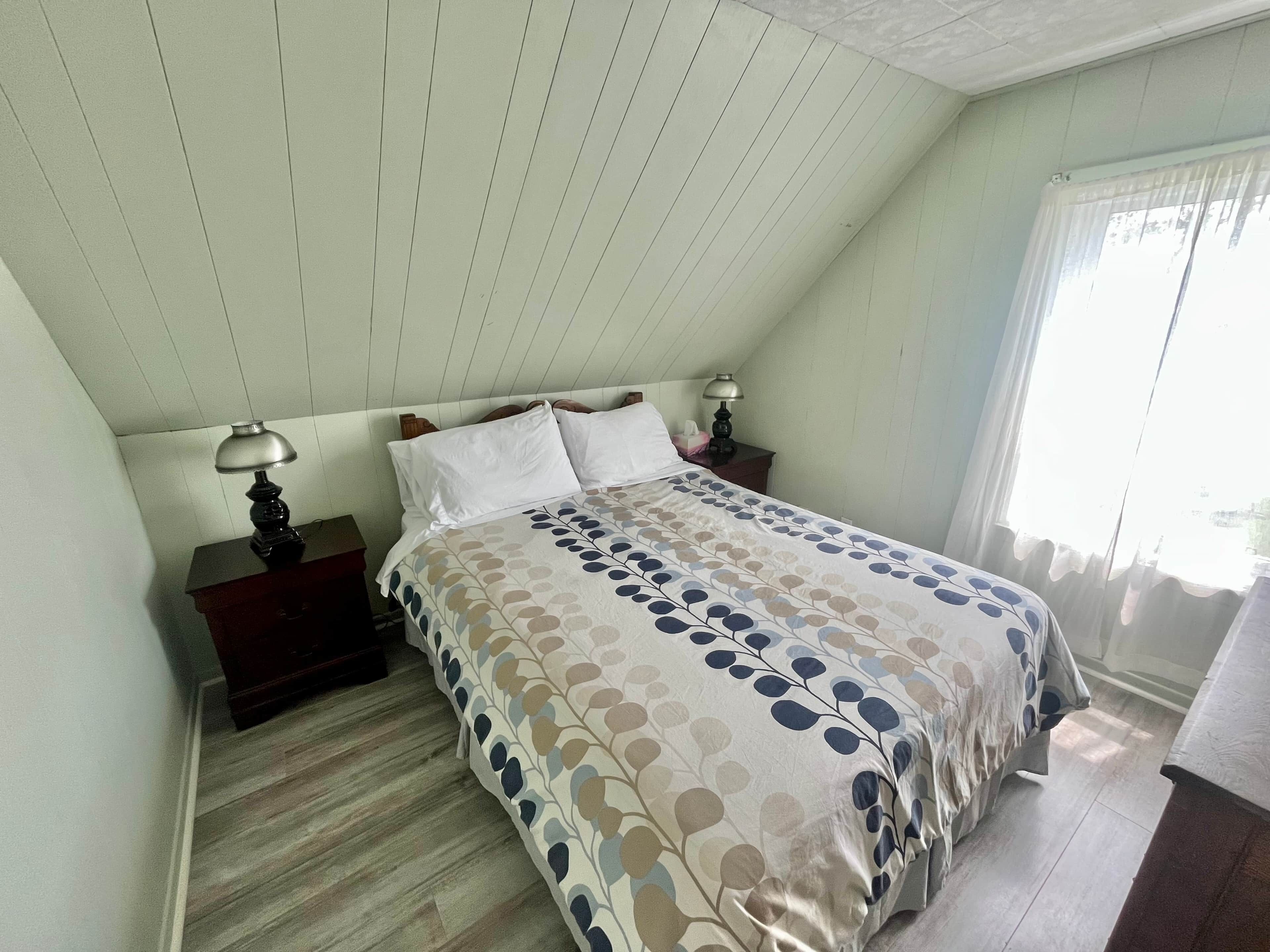 Une chambre avec un plafond en pente, un lit recouvert d’une couvre-lit à motifs, deux lampes de chevet et une fenêtre avec des rideaux.