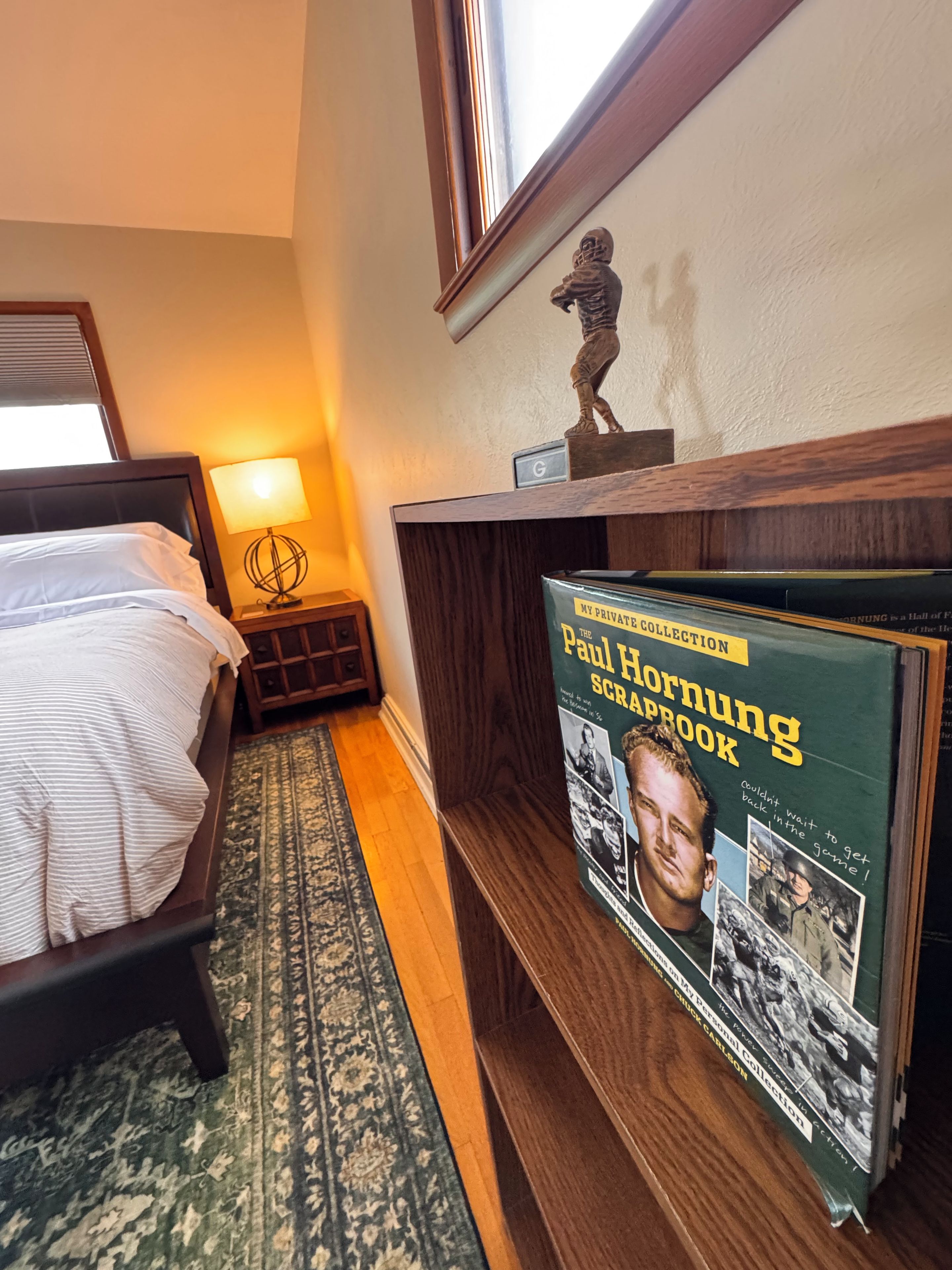 Ein Hotelzimmer mit einem Bett, einem Nachttisch mit Lampe, einer Holzablage mit dem Buch 'Paul Hornung Scrapbook' und einer dekorativen Statue.