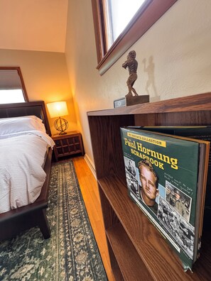 A hotel room with a bed, a nightstand with a lamp, a wooden shelf with a book titled 'Paul Hornung Scrapbook,' and a decorative statue. - Green Bay Original Across From Lambeau Field: Where Every Stay Becomes Tradition (Green Bay)