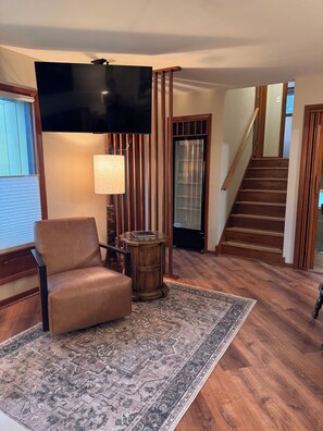 A living room with a brown leather chair, a patterned rug, a wooden side table, a flat-screen TV, and a staircase. - Green Bay Original Across From Lambeau Field: Where Every Stay Becomes Tradition (Green Bay)