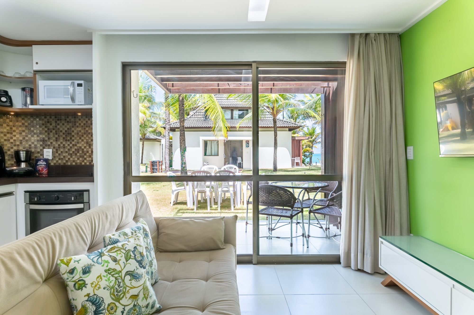 Uma sala de estar moderna com sofá, área de jantar com cadeiras, cozinha equipada e uma grande porta de vidro deslizante que dá para um pátio com mobiliário exterior e vista para um edifício com telhado telhado.