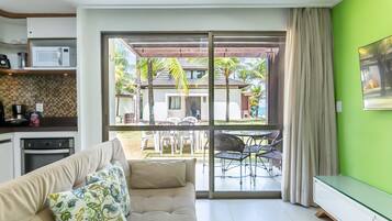 Uma sala de estar moderna com sofá, área de jantar com cadeiras, cozinha equipada e uma grande porta de vidro deslizante que dá para um pátio com mobiliário exterior e vista para um edifício com telhado telhado.