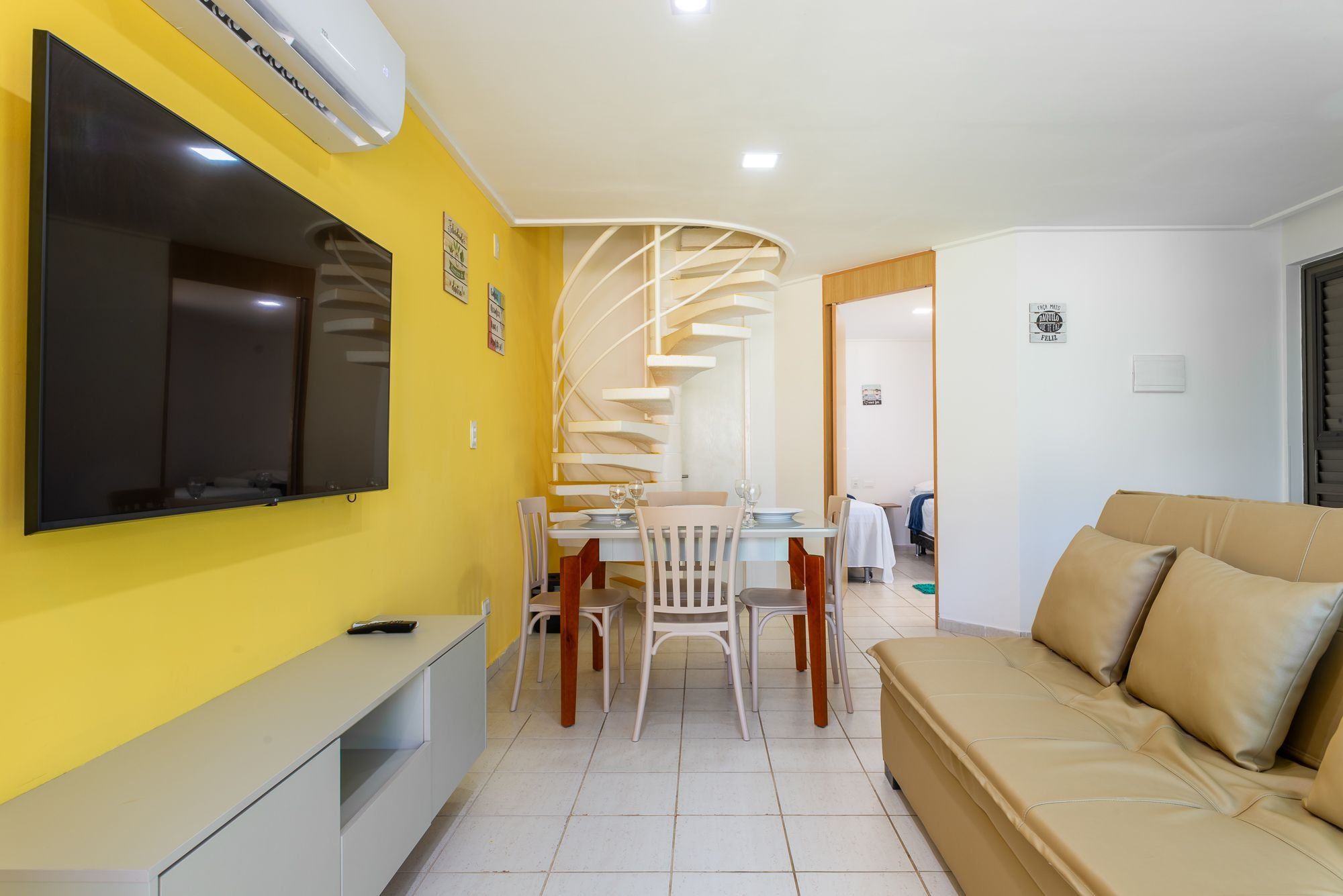 Uma sala de estar moderna com sofá, televisão fixada na parede, área de jantar com cadeiras e mesa, e uma escada em espiral que leva a outro cômodo.