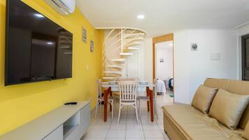 Uma sala de estar moderna com sofá, televisão fixada na parede, área de jantar com cadeiras e mesa, e uma escada em espiral que leva a outro cômodo.