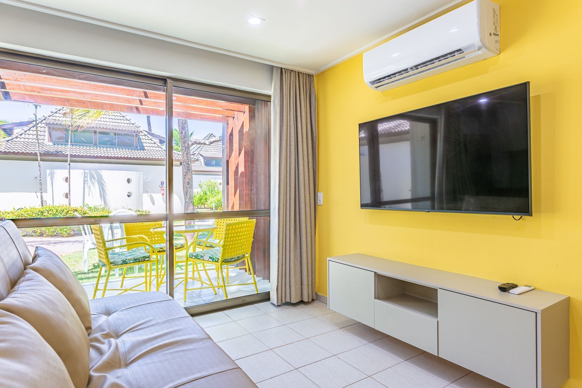 Una sala de estar moderna con un sofá, un televisor de pantalla plana montado en la pared, una puerta corrediza de vidrio que da a un área exterior con sillas amarillas y un mueble blanco.
