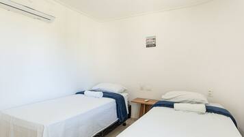 A small, clean room with two single beds, a bedside table with a lamp, and a wall-mounted air conditioning unit.
