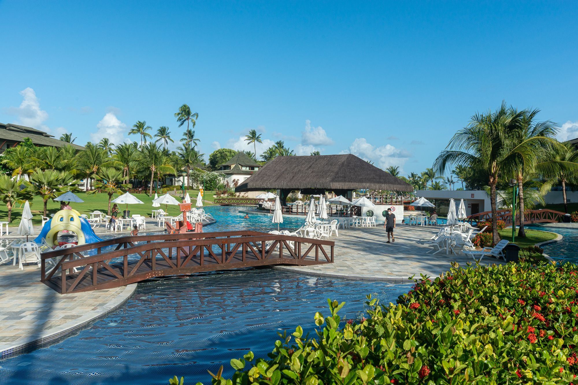 Área de piscina do resort com ponte, espreguiçadeiras, guarda-sóis e palmeiras.