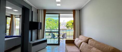 Una sala de estar moderna con sofá, televisor de pantalla plana, mesa de vidrio y un balcón con vista a la vegetación.