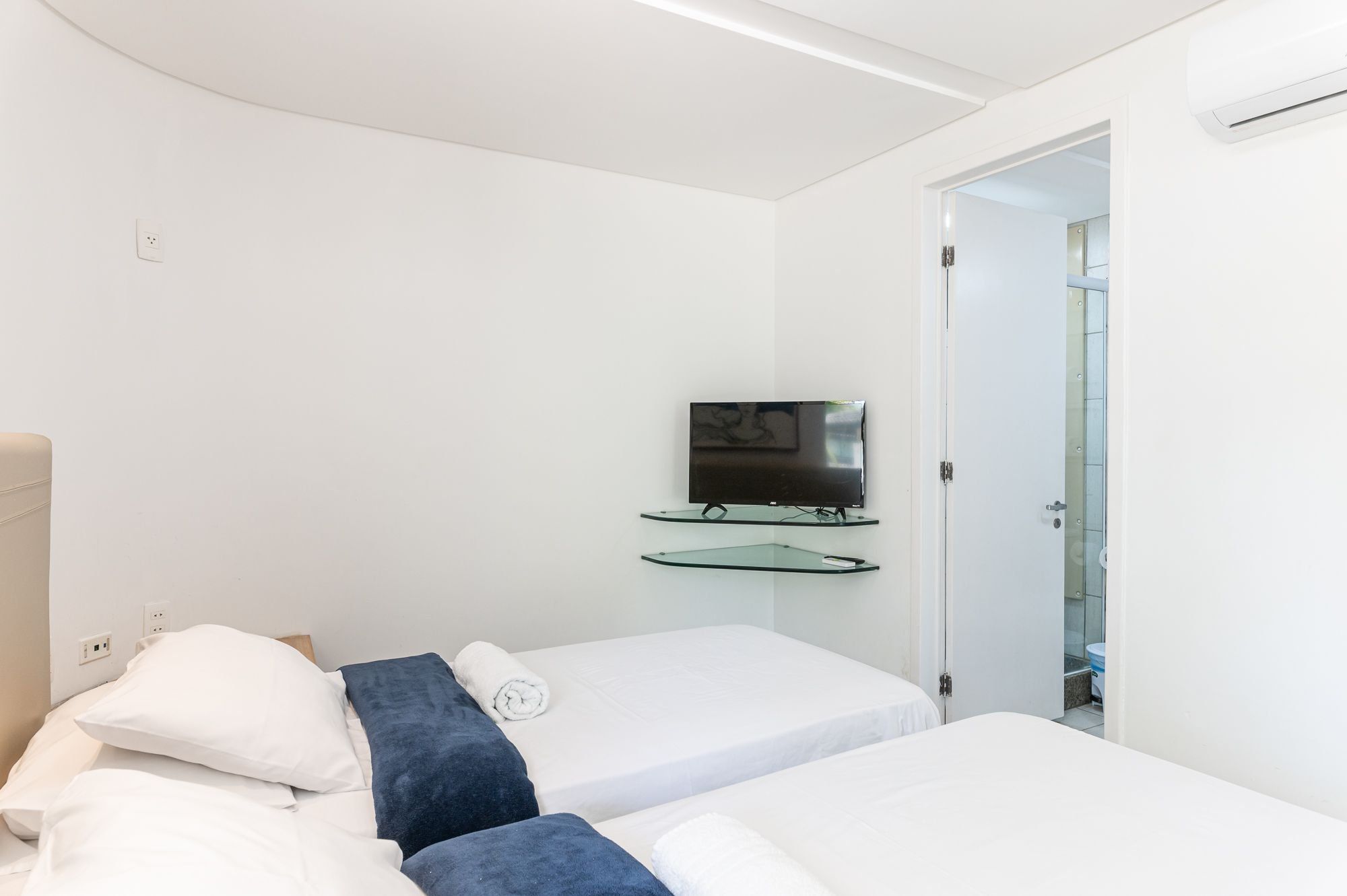 A hotel room with two beds, a television mounted on a glass shelf, and a door leading to a bathroom.