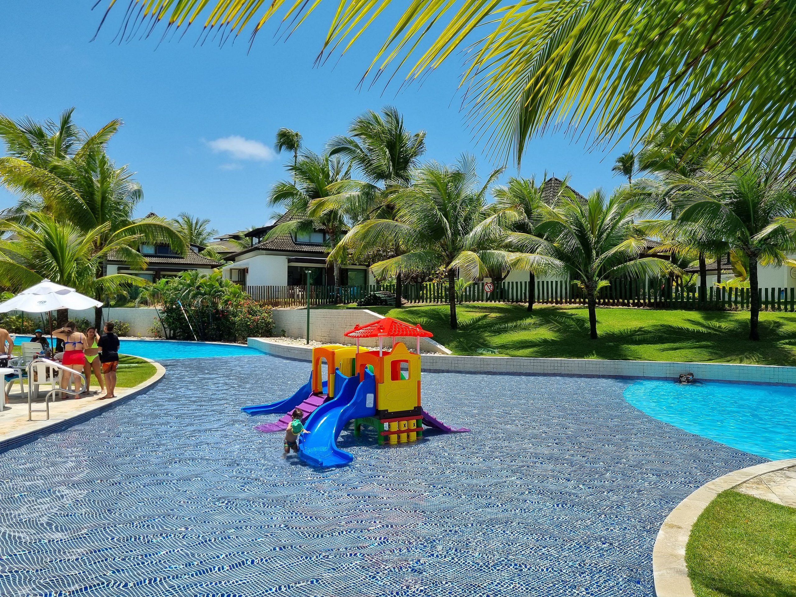 A swimming pool area with a children's play structure, palm trees, and a fenced residential area in the background.