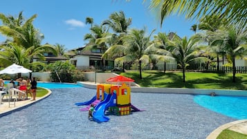 A swimming pool area with a children's play structure, palm trees, and a fenced residential area in the background.