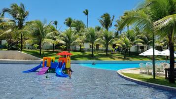 Área de piscina com brinquedos infantis, palmeiras e espreguiçadeiras.