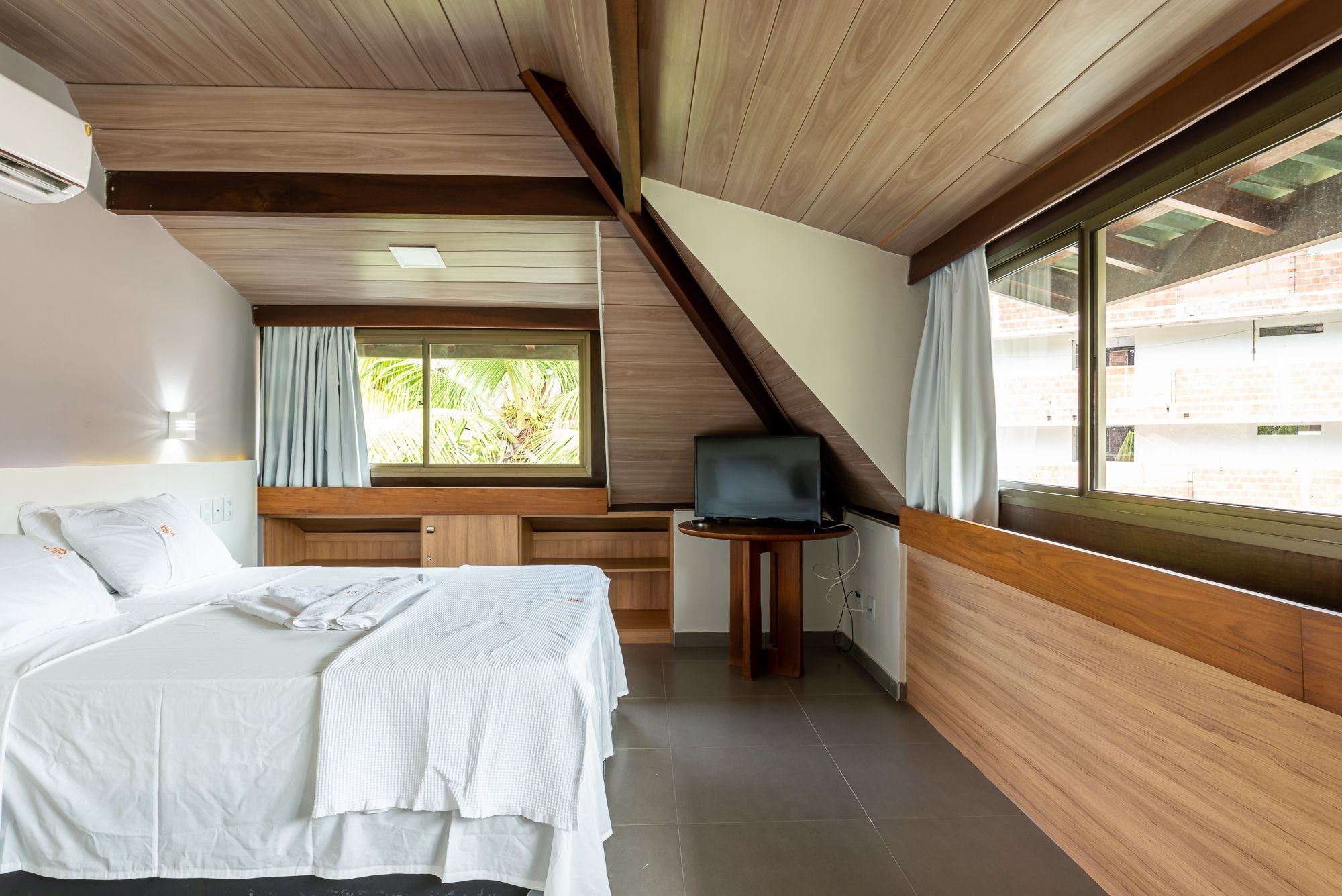Um quarto com cama, televisão sobre uma mesinha, janela com cortinas e teto com vigas de madeira.