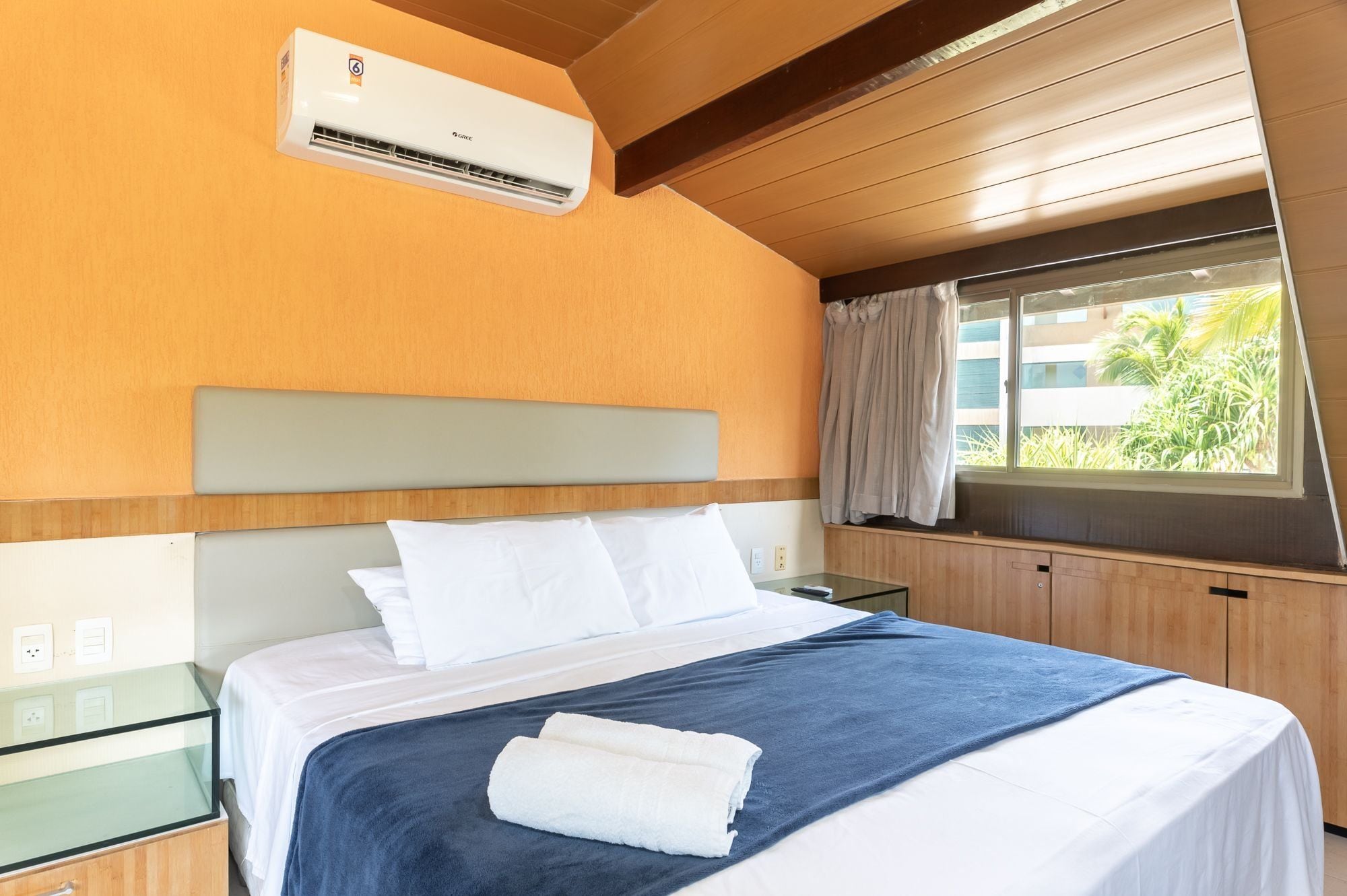 Quarto de hotel com uma cama, uma mesa de cabeceira de vidro, um criado-mudo e uma janela com cortinas, além de um aparelho de ar condicionado.