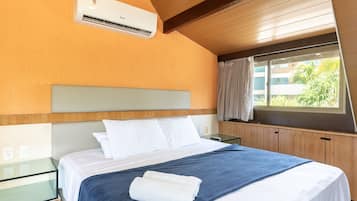 Quarto de hotel com uma cama, uma mesa de cabeceira de vidro, um criado-mudo e uma janela com cortinas, além de um aparelho de ar condicionado.