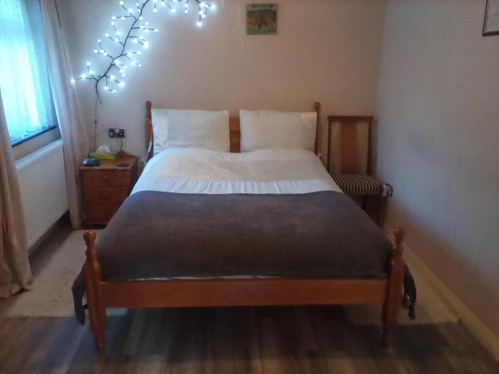 A bedroom with a wooden bed, white bedding, a nightstand with a lamp, and a wall-mounted light fixture.