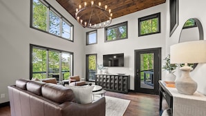 A spacious living room with a leather sofa, a dining area, a fireplace, and large windows offering views of trees.