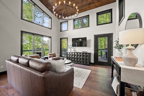 A spacious living room with a leather sofa, a dining area, a fireplace, and large windows offering views of trees.