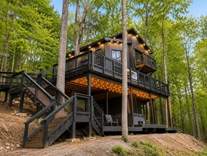 A multi-level cabin with outdoor seating and string lights, surrounded by trees.