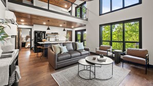 A spacious living area with a high ceiling, a sofa, a coffee table, and large windows offering a view of greenery.